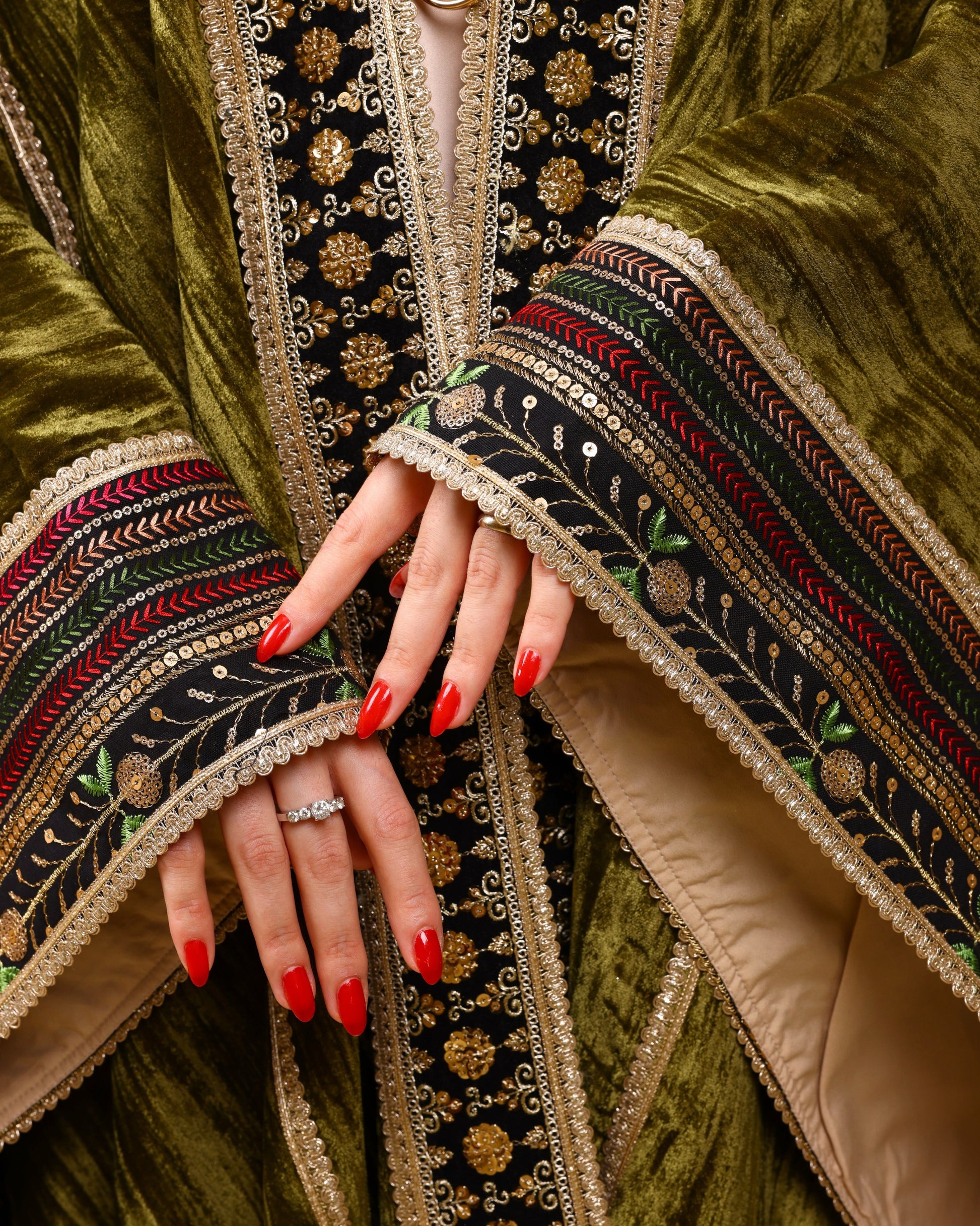 Close-up of a hand with red nail polish wearing a ring, against the backdrop of an intricately embroidered garment.