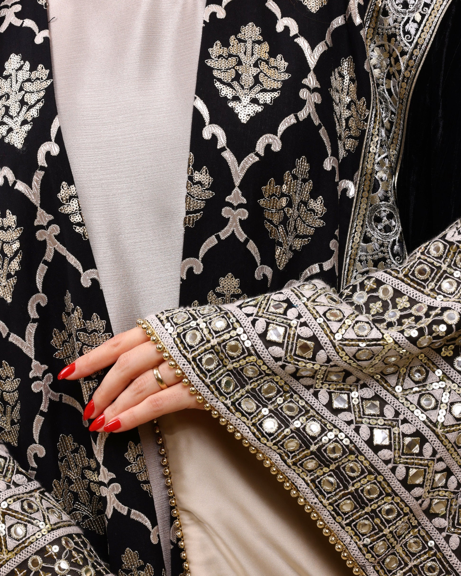 Close-up of an intricately designed black garment with silver embroidery, featuring a hand with red nail polish.