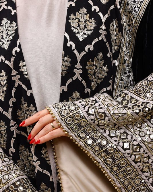 Close-up of an intricately designed black garment with silver embroidery, featuring a hand with red nail polish.