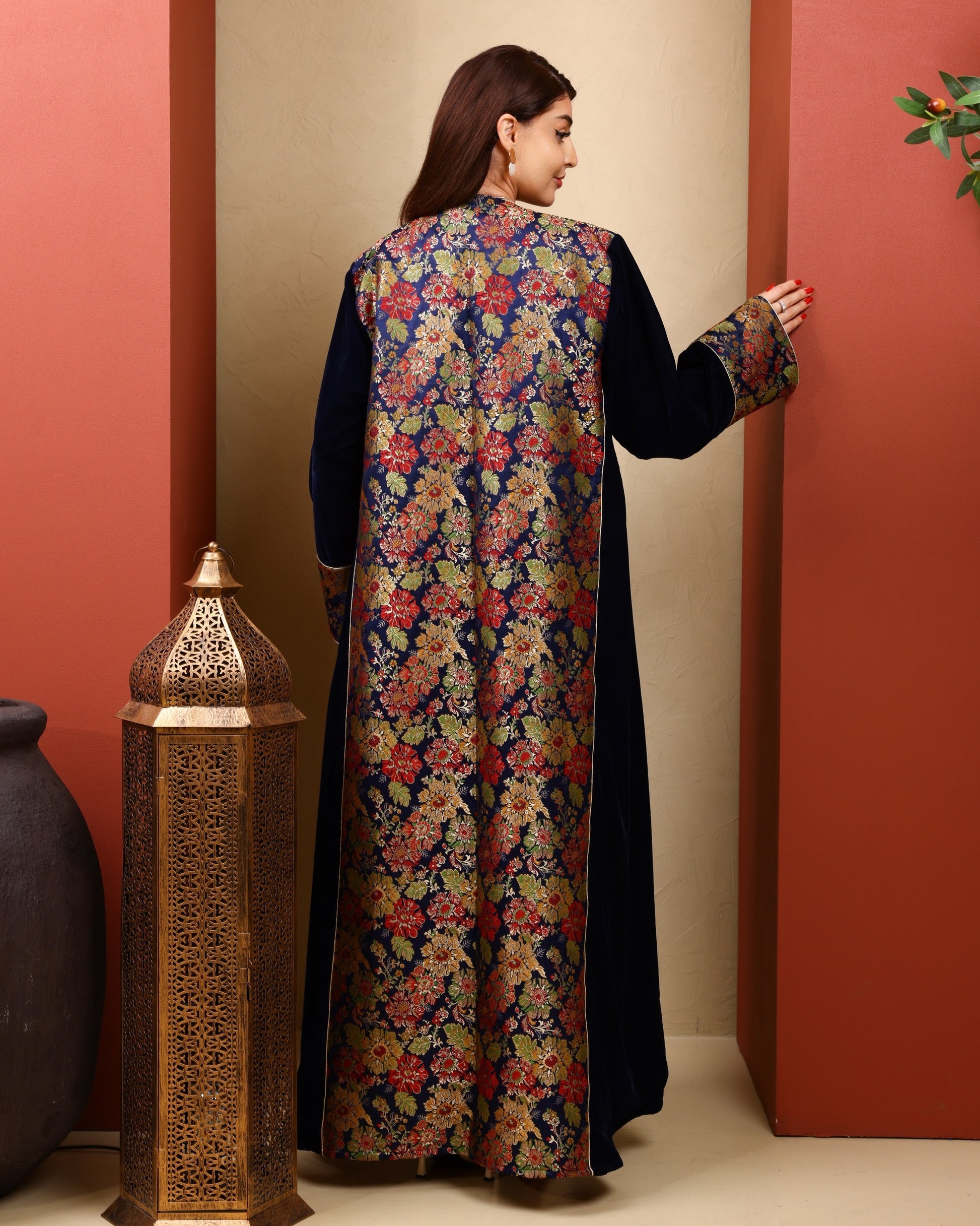 Woman wearing a long, dark-colored dress with a colorful floral pattern, standing against a warm-toned wall.