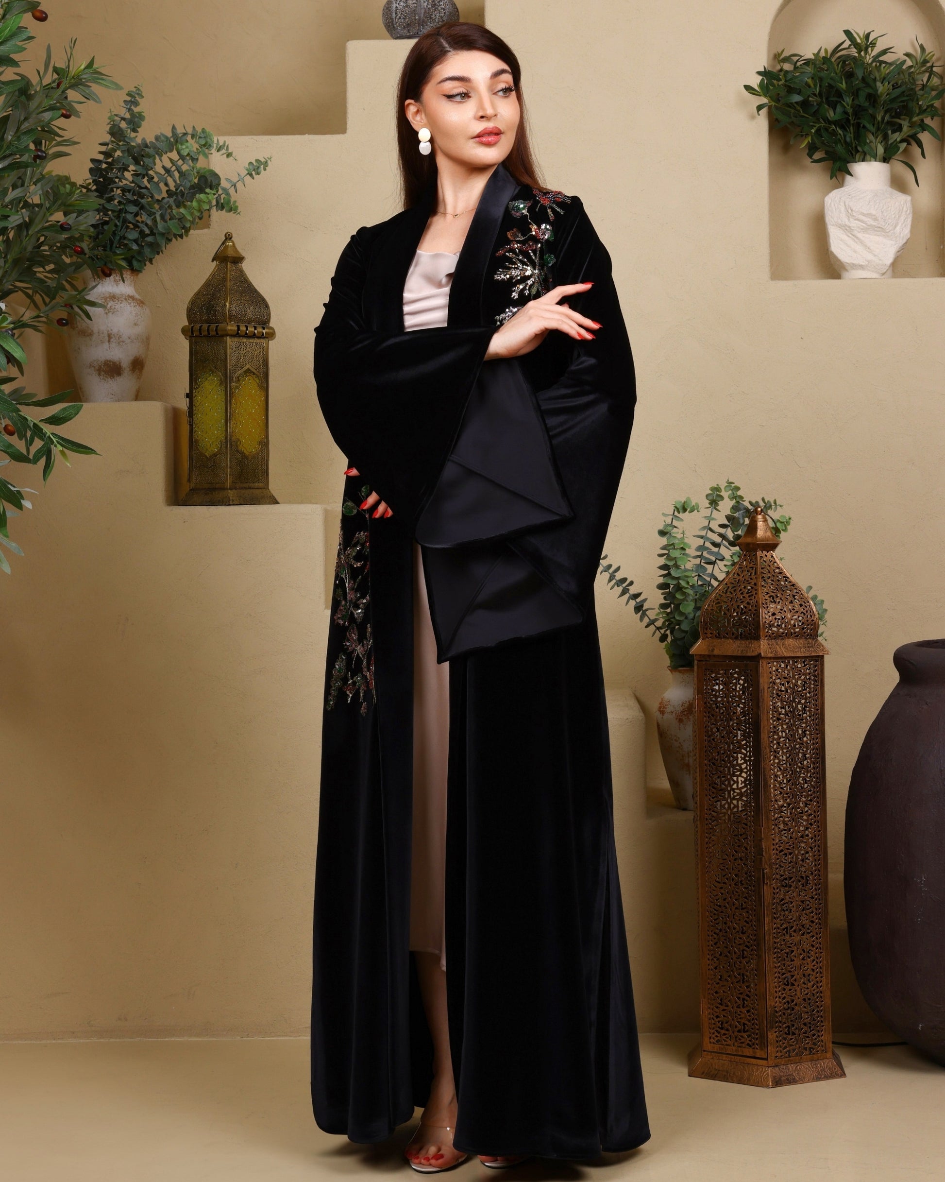 Woman wearing a black embroidered abaya in a decorated room with plants and lanterns.