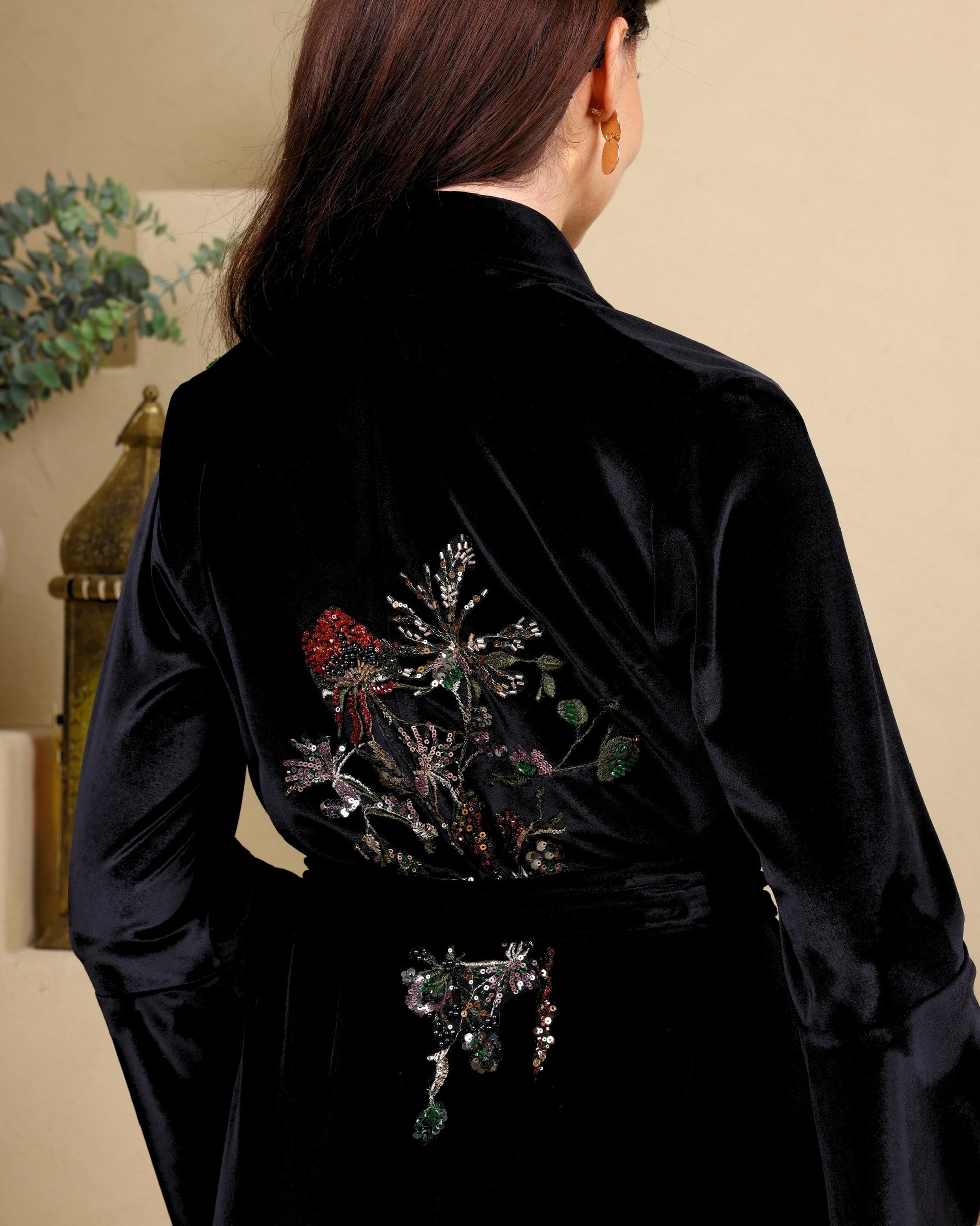 Person wearing a black robe with floral embroidery on the back, standing against a beige wall.