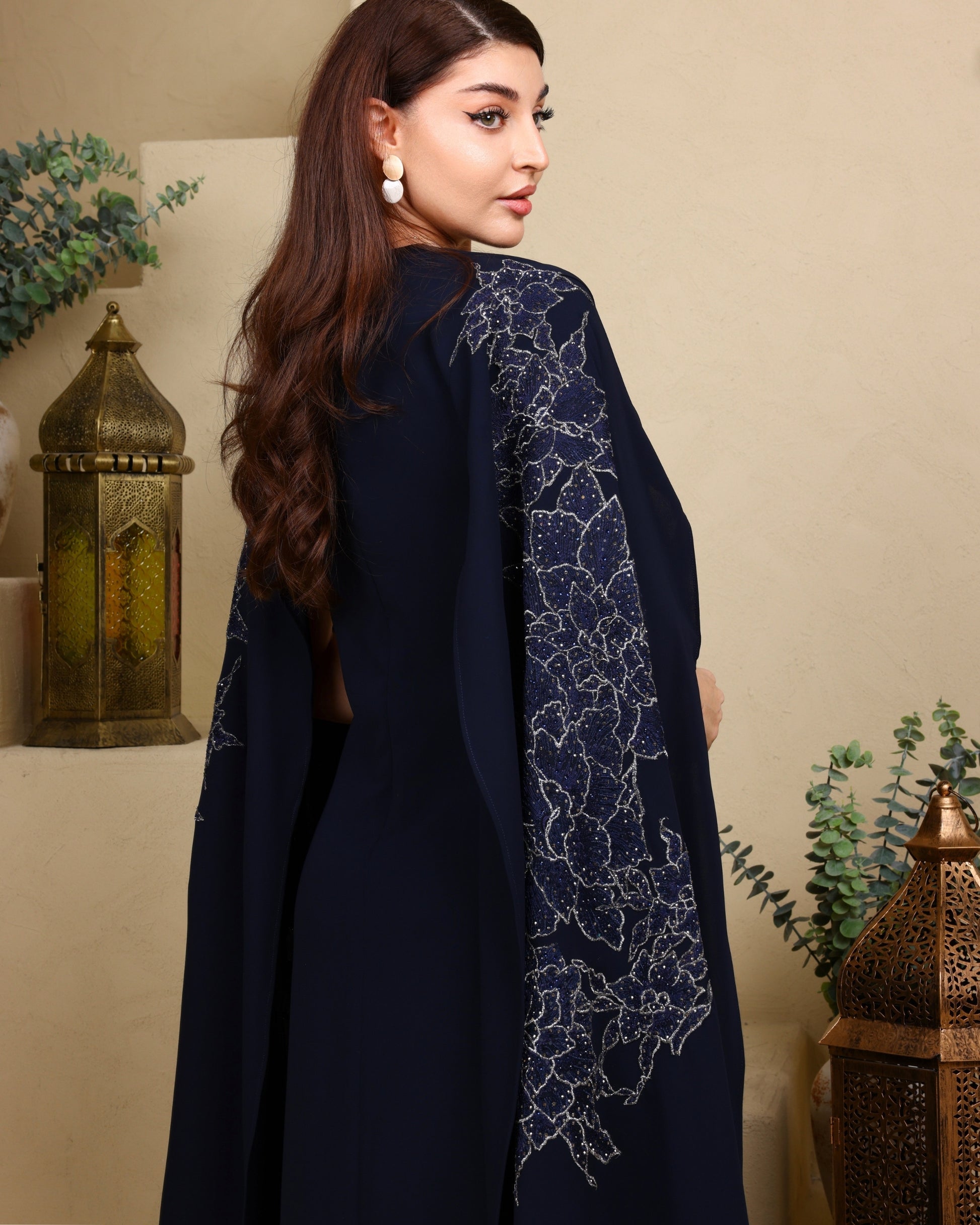 Woman wearing a dark blue outfit with intricate sequin embroidery, standing against a beige wall with decorative elements.