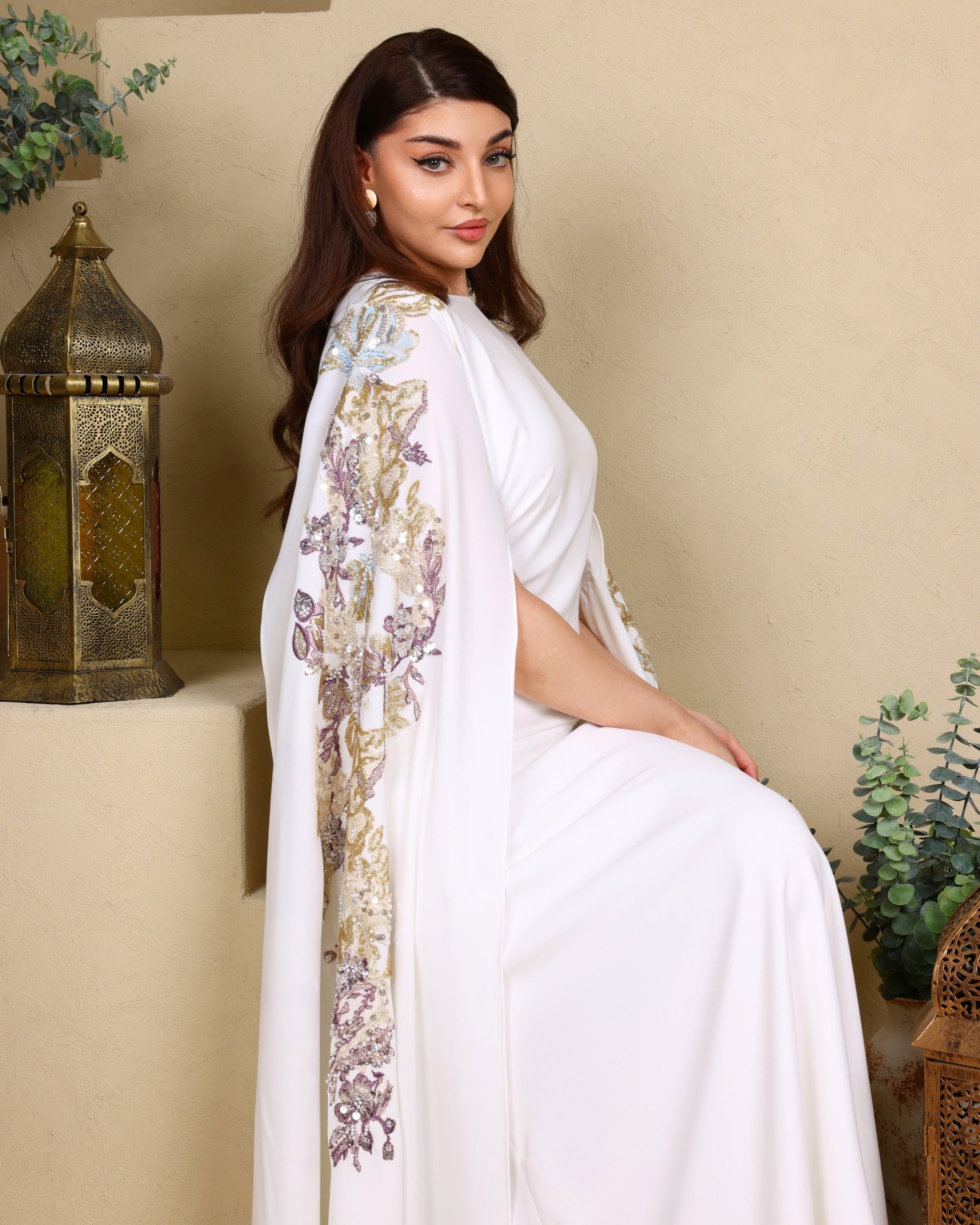 Woman in a white saree with floral patterns sitting against a beige wall.