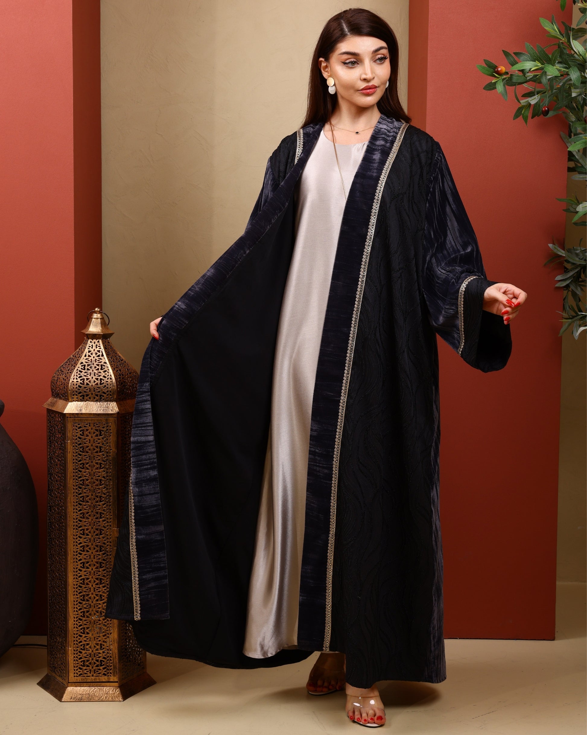 Woman wearing a long dark grey abaya with a white inner garment, standing against a red wall.