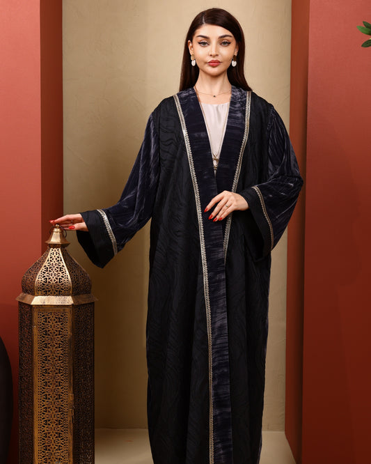 Woman wearing a black abaya with gold trim against a warm-toned wall.