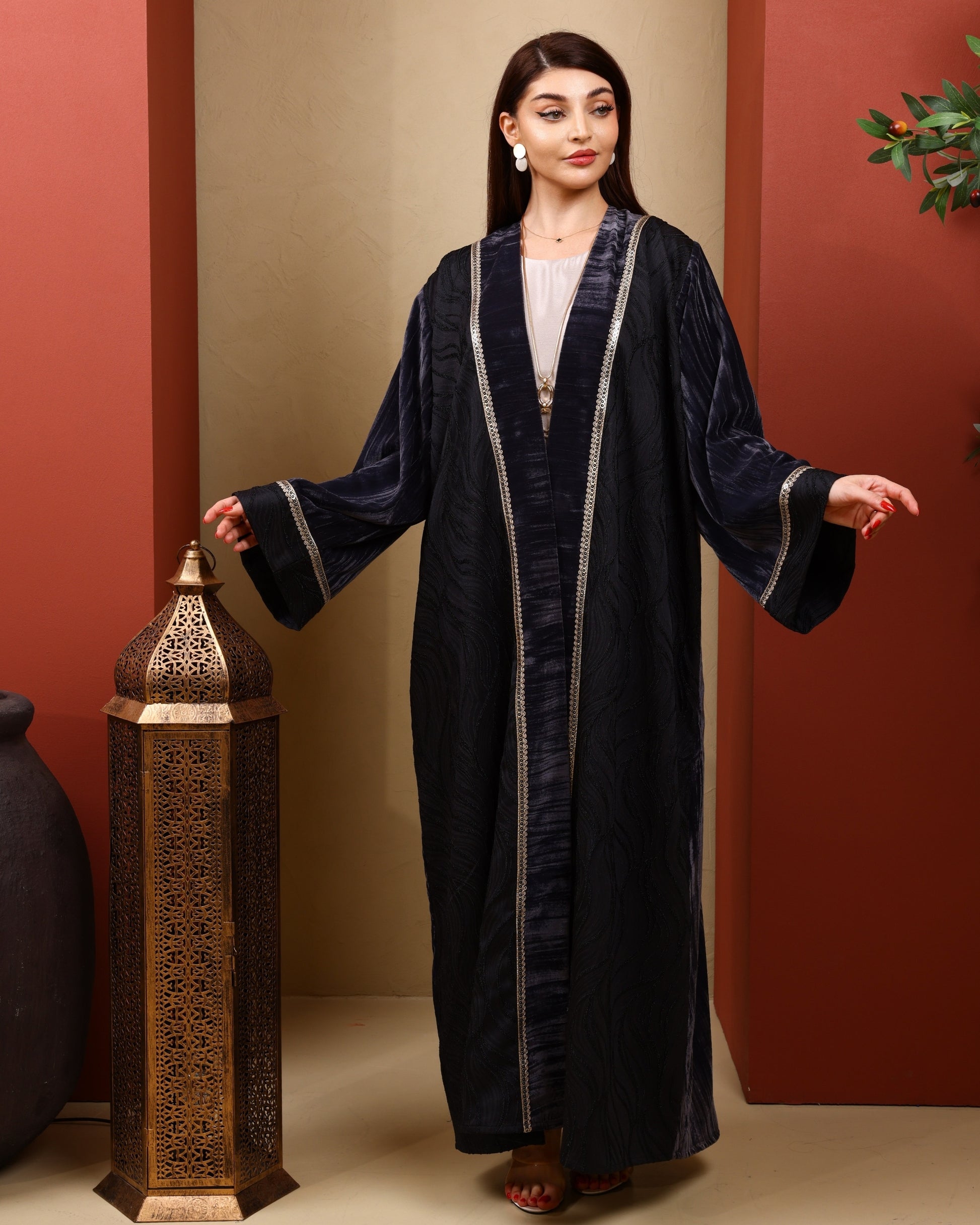 Woman wearing a long dark grey abaya with gold trim against a warm-toned wall.
