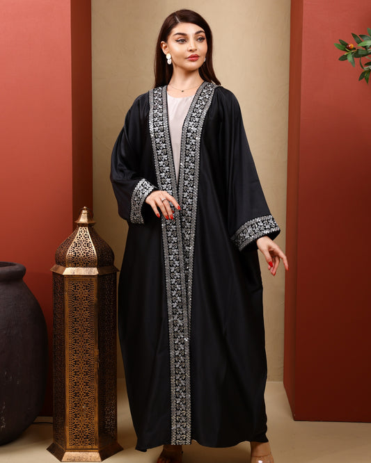 Woman wearing a black abaya with intricate silver embroidery in a warm-toned room.