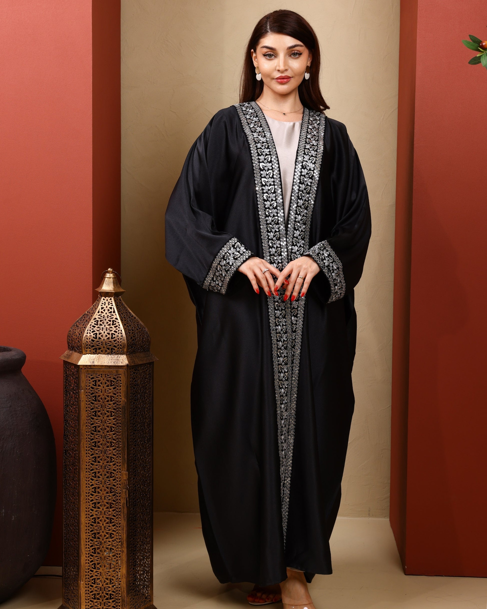 Woman wearing a black abaya with silver embroidery against a beige wall.