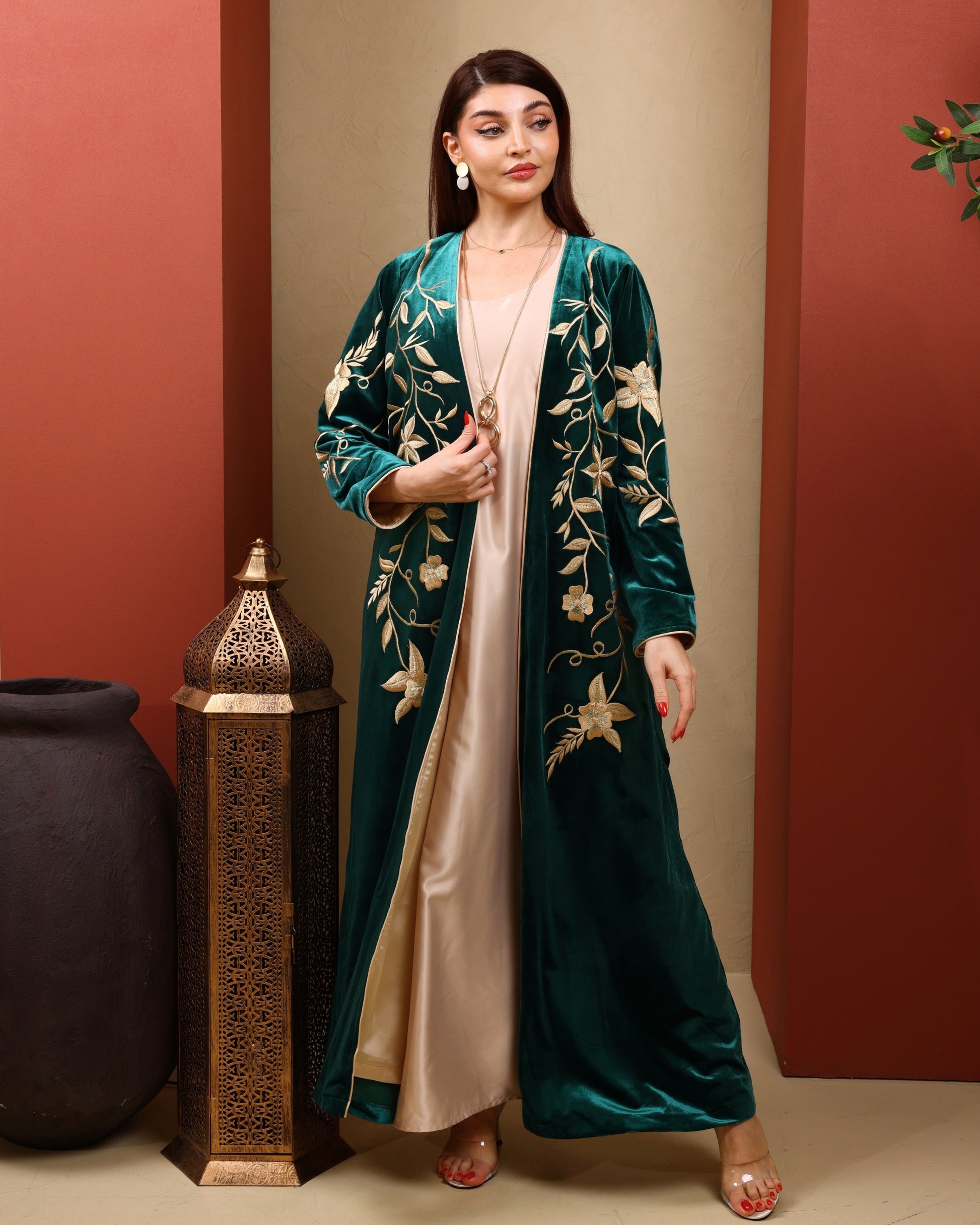 Woman wearing a green embroidered abaya with a beige dress underneath, standing against a warm-toned wall.