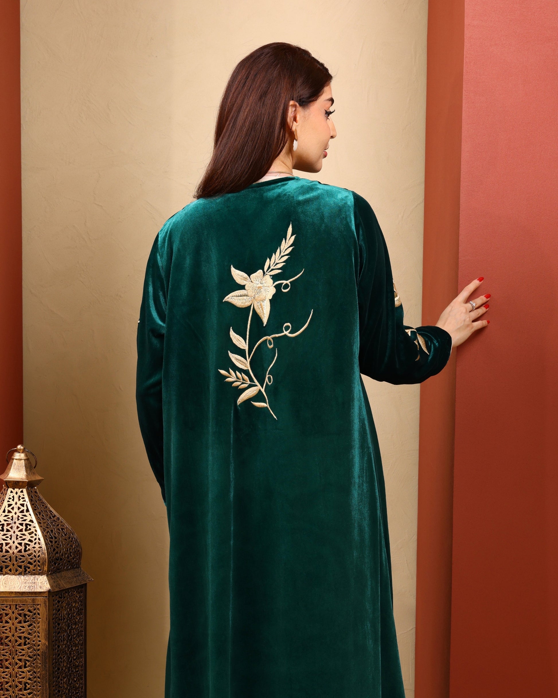 Woman wearing a green velvet dress with floral embroidery, standing against a beige and red wall.