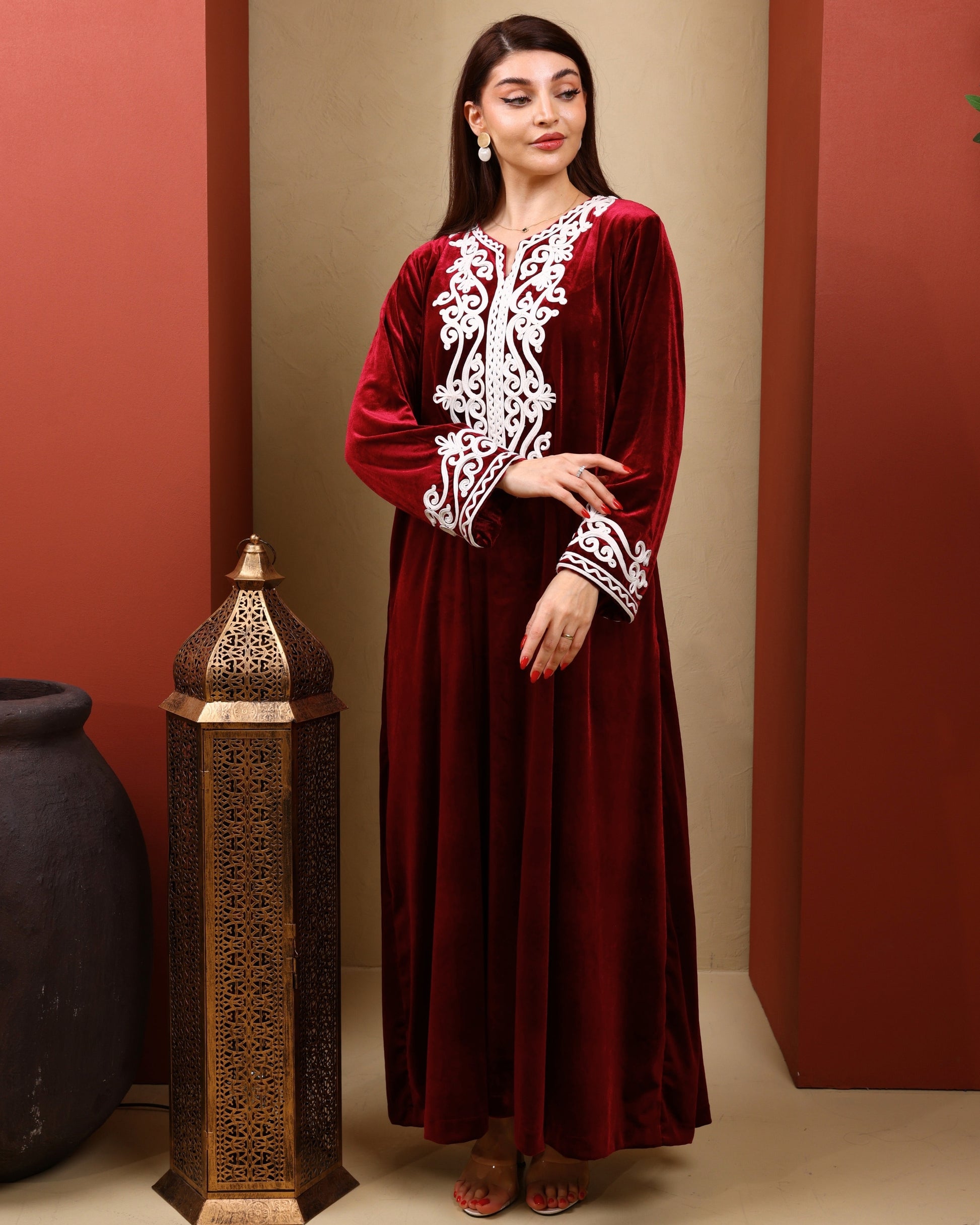 Woman wearing a red velvet dress with white embroidery against a warm-toned wall.