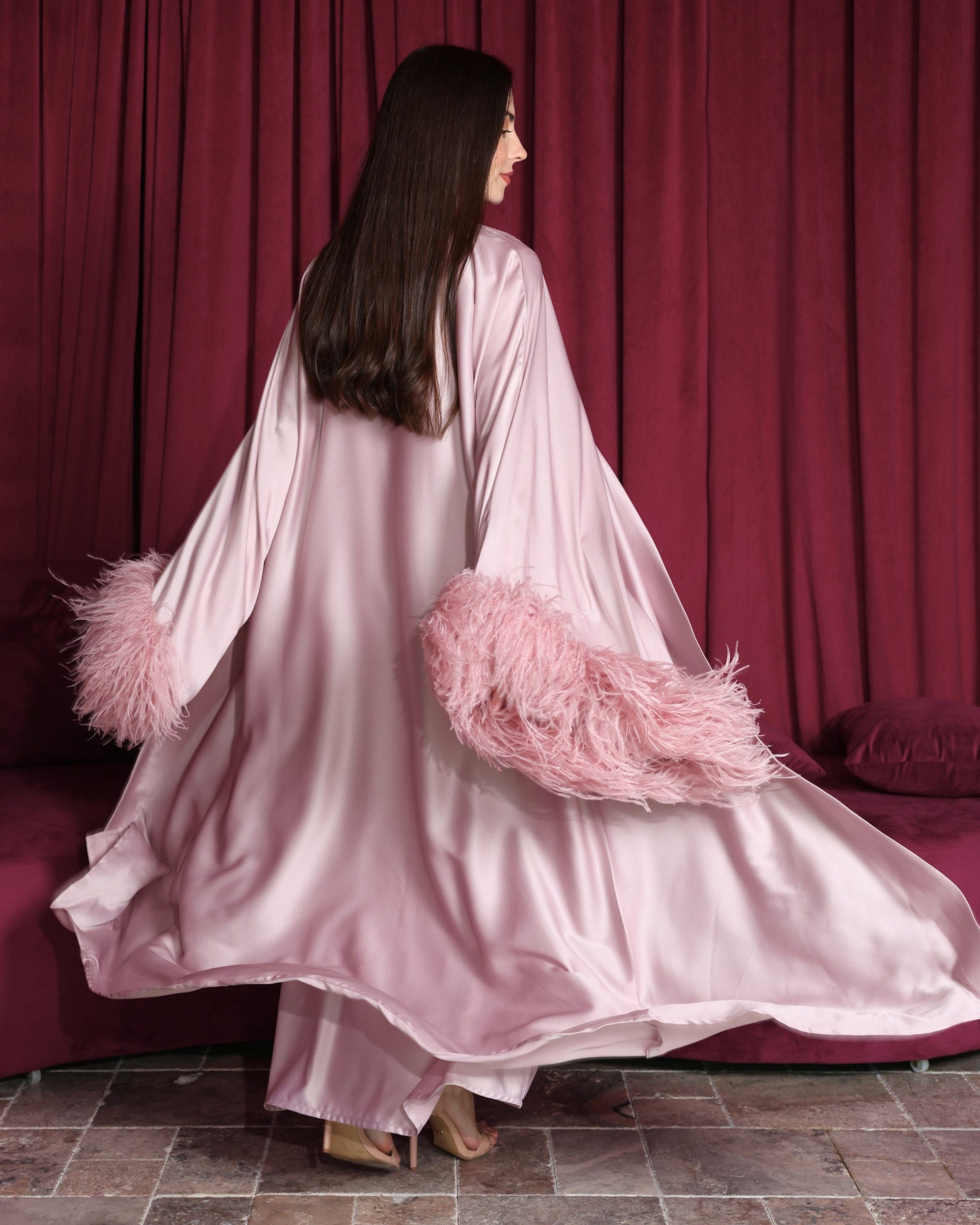 Woman wearing a pink satin robe with feathered sleeves against a red curtain background