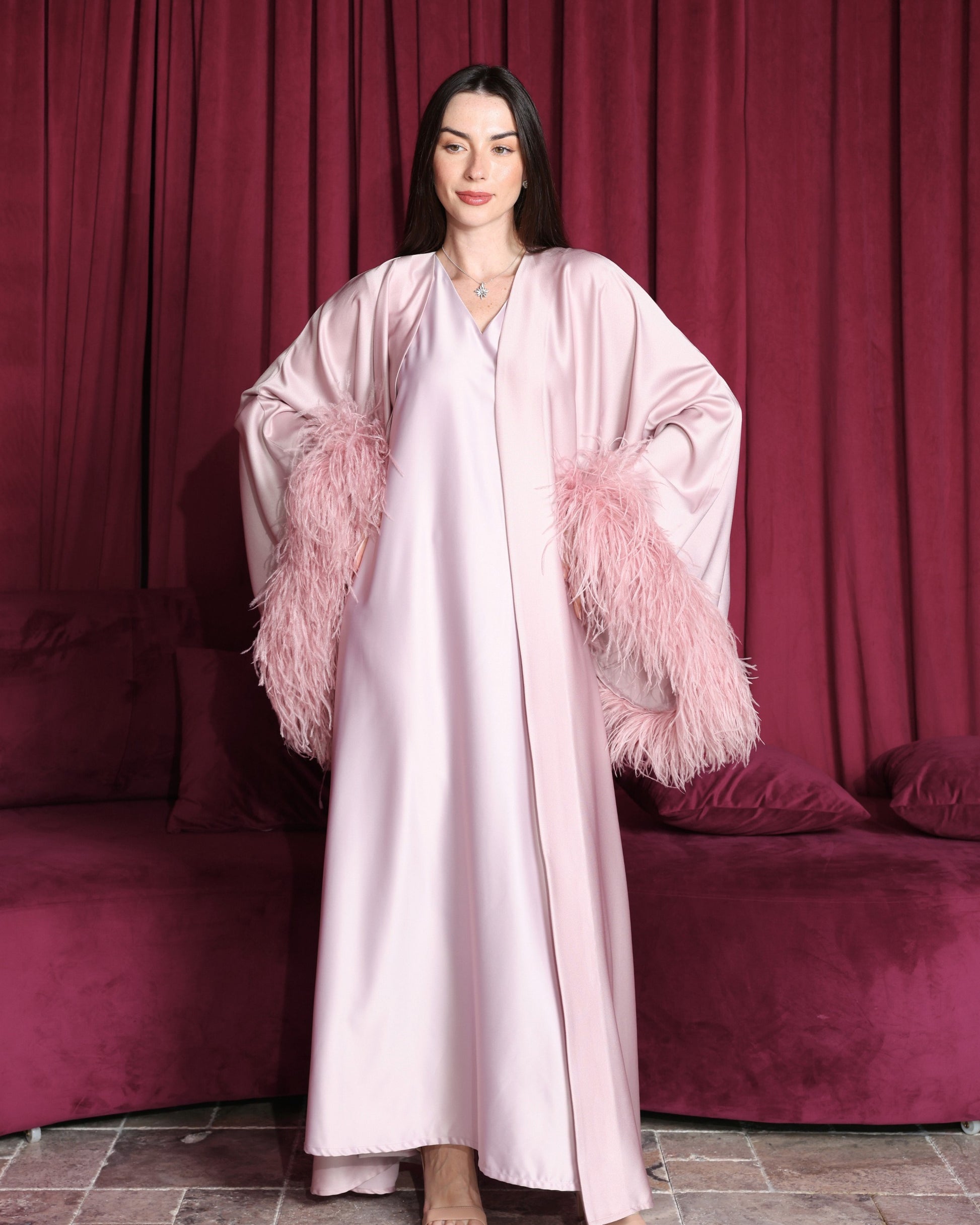 Woman wearing a light pink robe with feathered sleeves against a red curtain backdrop